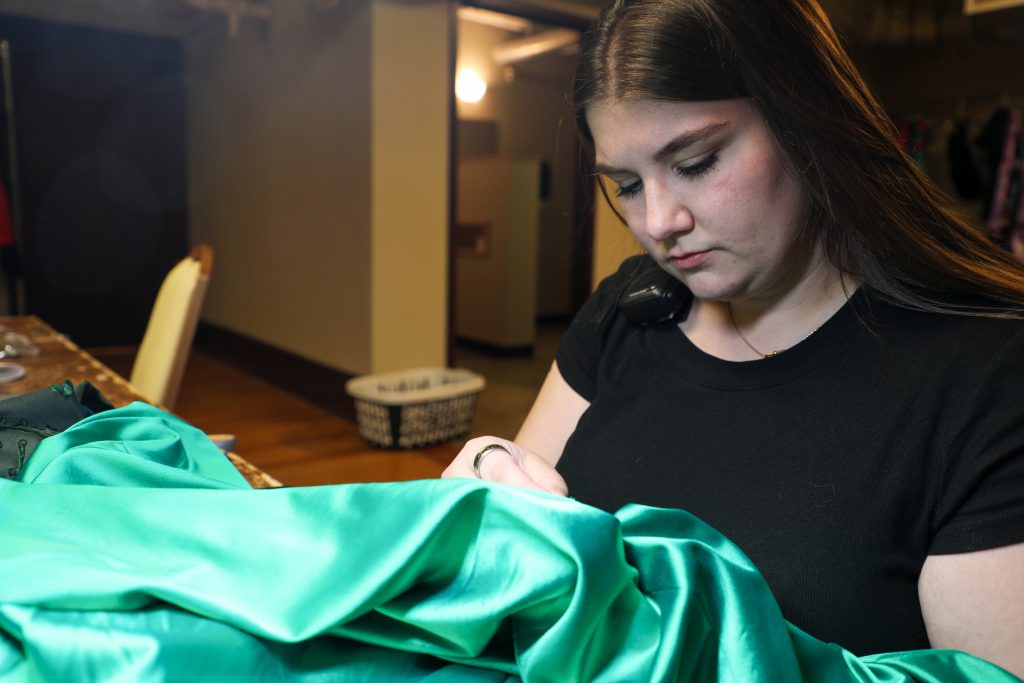 A young woman works intently, sewing repairs on a green dress