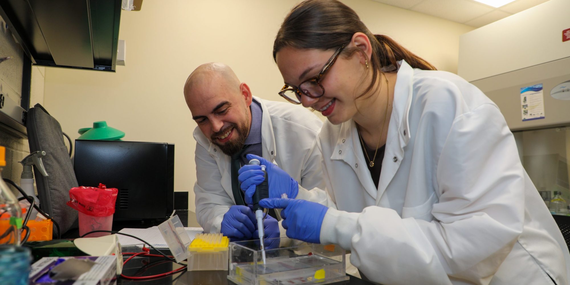 An RMC student uses a pipette inside a biology lab under observation of a professor