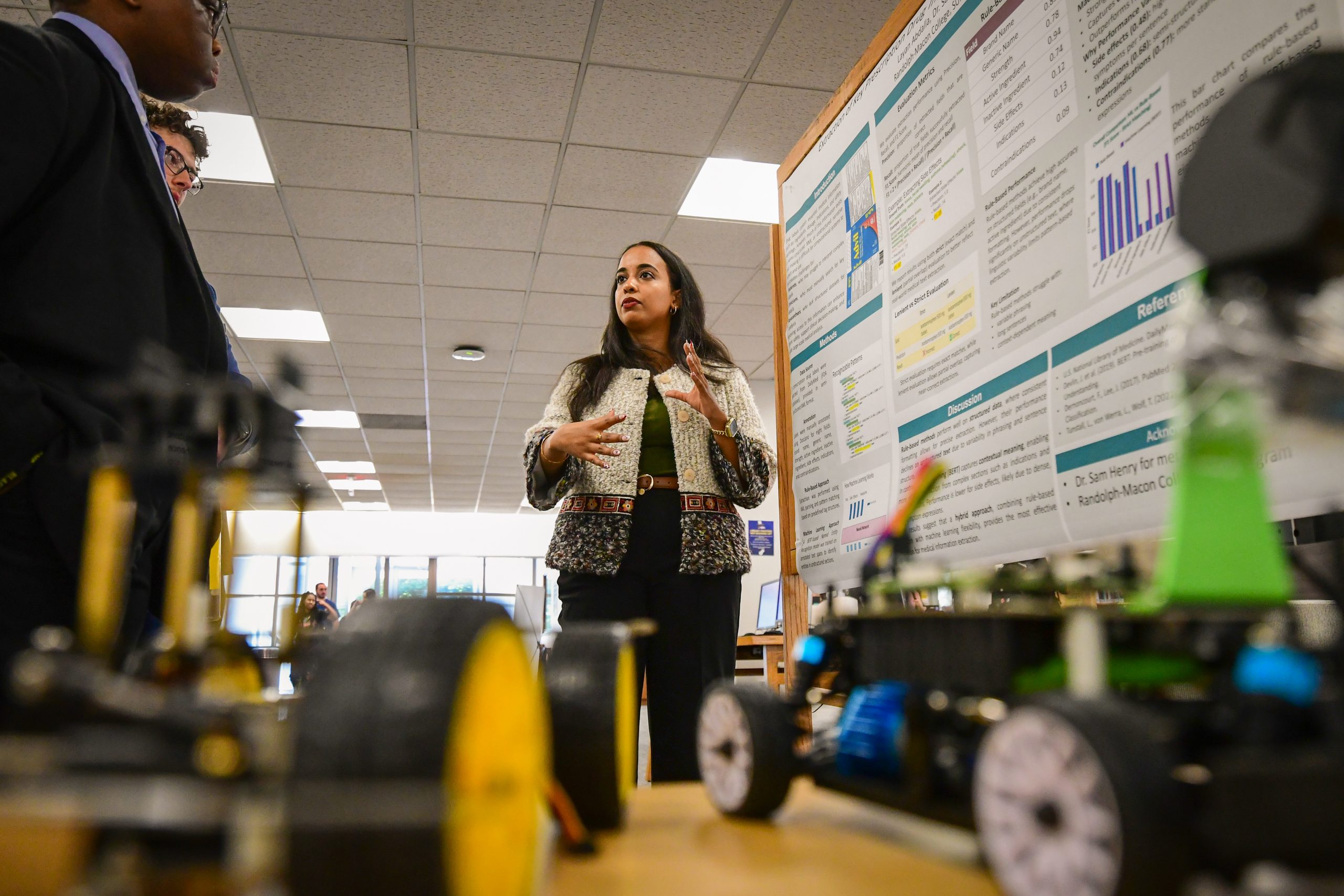 With robotic vehicles in the foreground, a student explains her research poster to attendees of a symposium