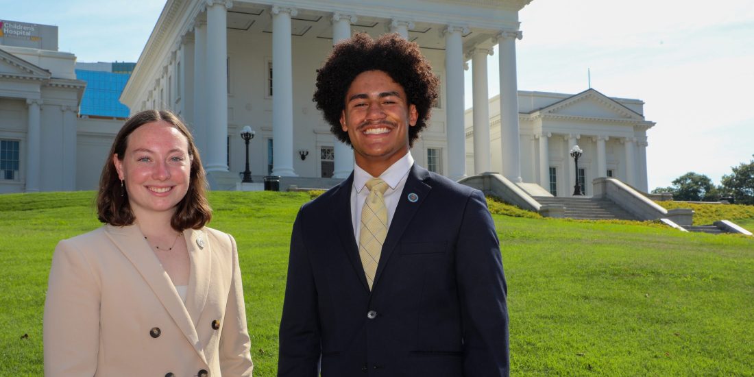 Two RMC students in business suits stand smiling in front of the Virginia Capitol building