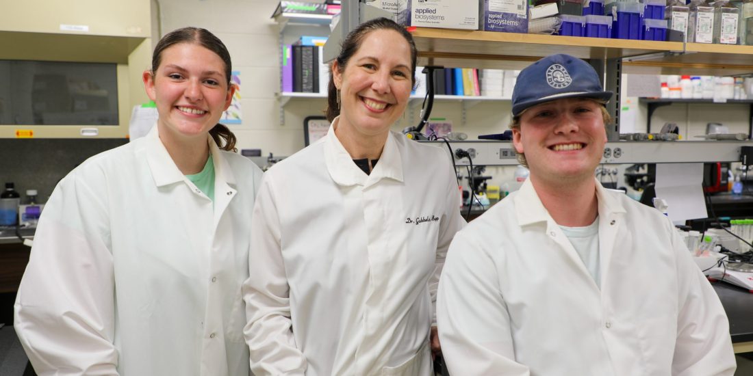 Two students in lab coats stand alongside a professor, all smiling, inside a Biology lab