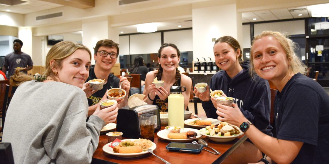 Students seated around a table inside Estes Dining Hall hold up bowls of food and smile for the camera
