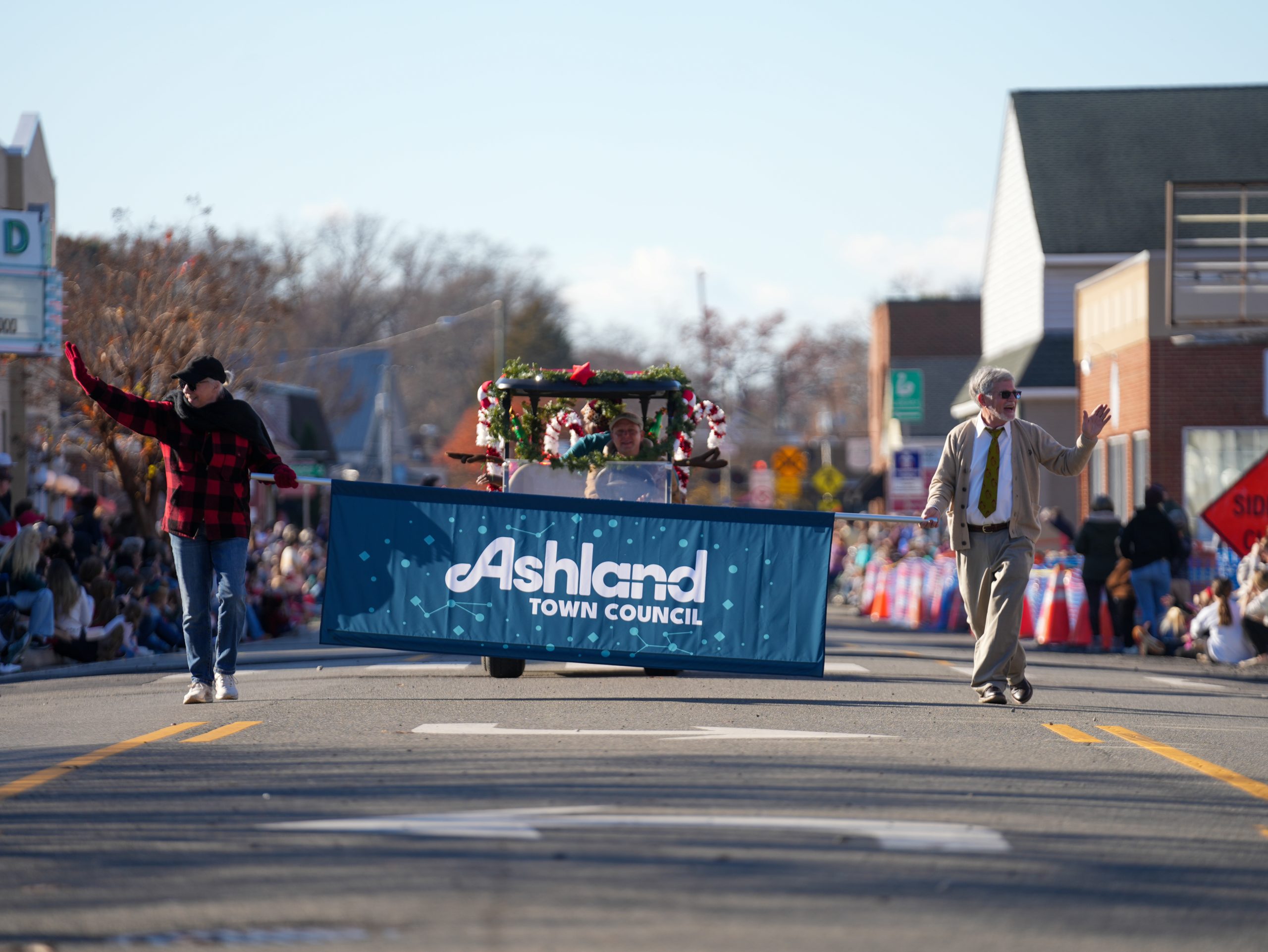 People including the Mayor of Ashland walk with a banner reading "Ashland" ahead of vehicle at the start of the Ashland Olde Time Holiday Parade.