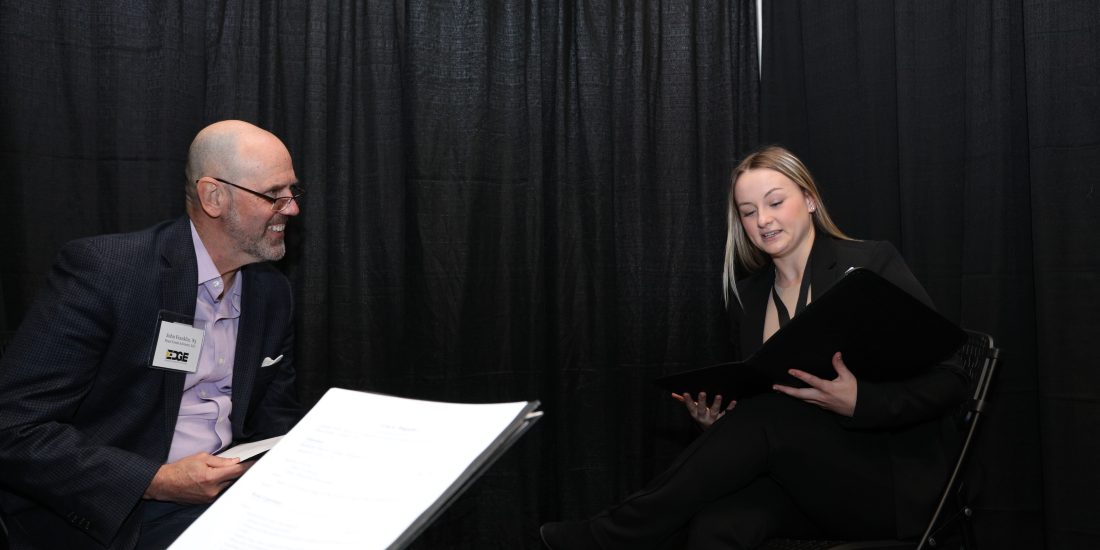 A paper and knee in the foreground with two people mid interview in the background