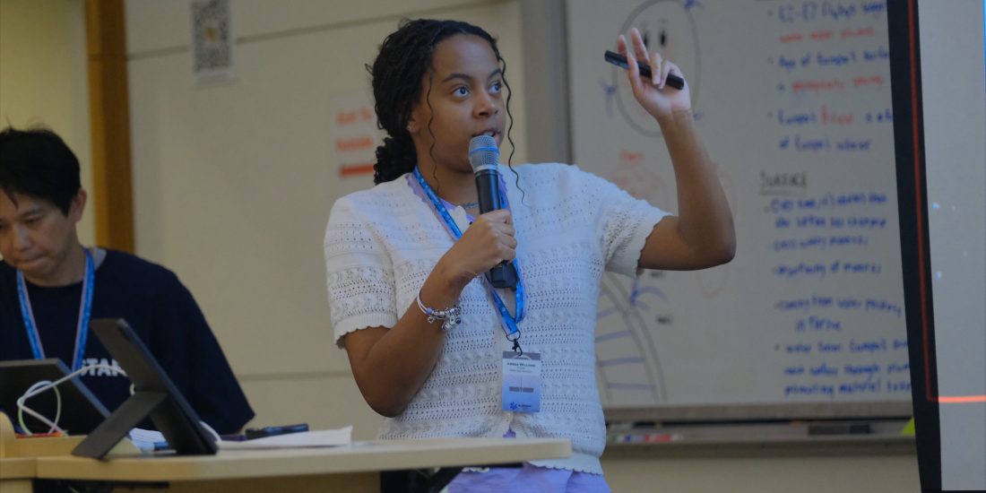 A student speaks into a microphone while presenting research in front of a classroom