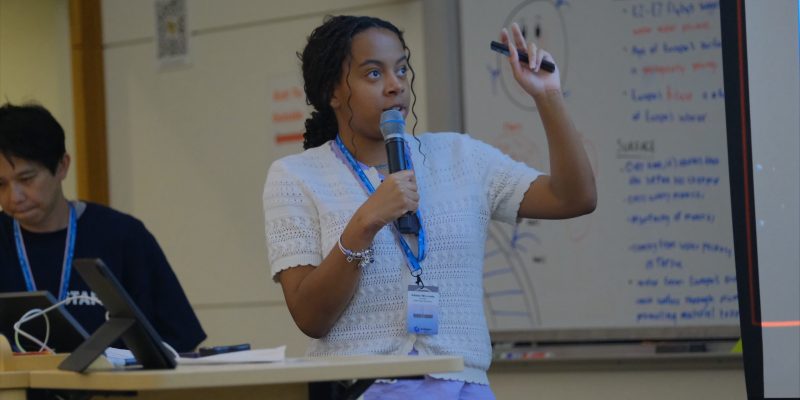 A student speaks into a microphone while presenting research in front of a classroom