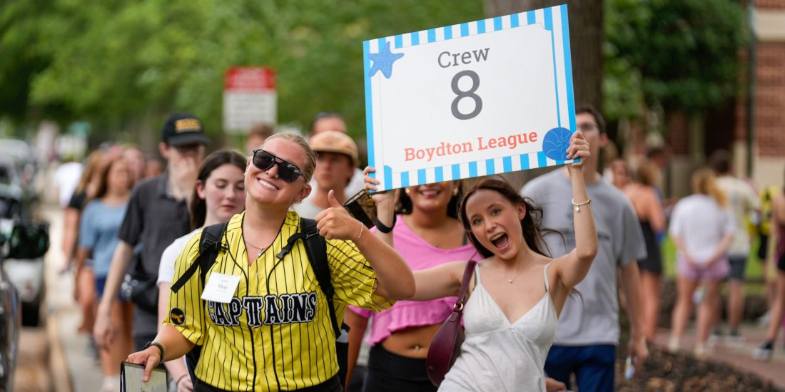 A student smiles and holds up a "Crew 8" sign while walking next to a Captain, who is holding a thumbs up sign and leading a group through campus