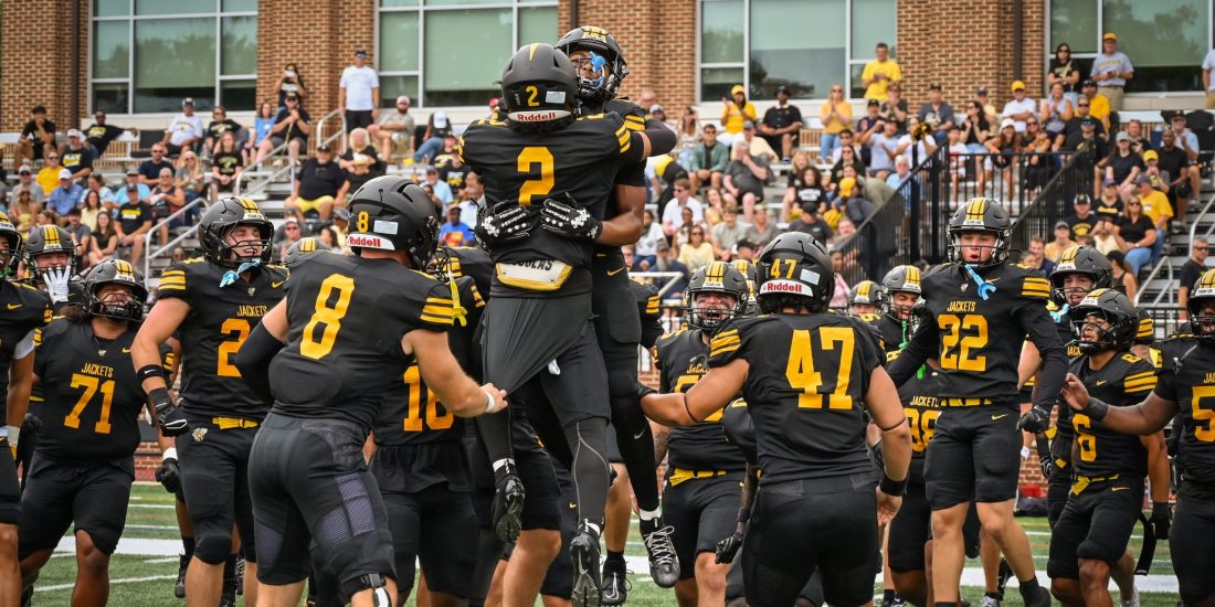 RMC football players jump and embrace in celebration during a game at Day Field in front of a packed crowd