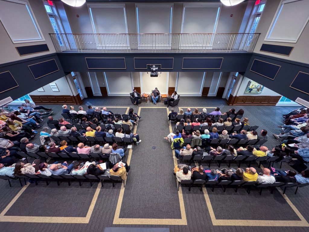 Several rows of attendees seated in chairs listen to three panelists at the front of a room in Birdsong Hall