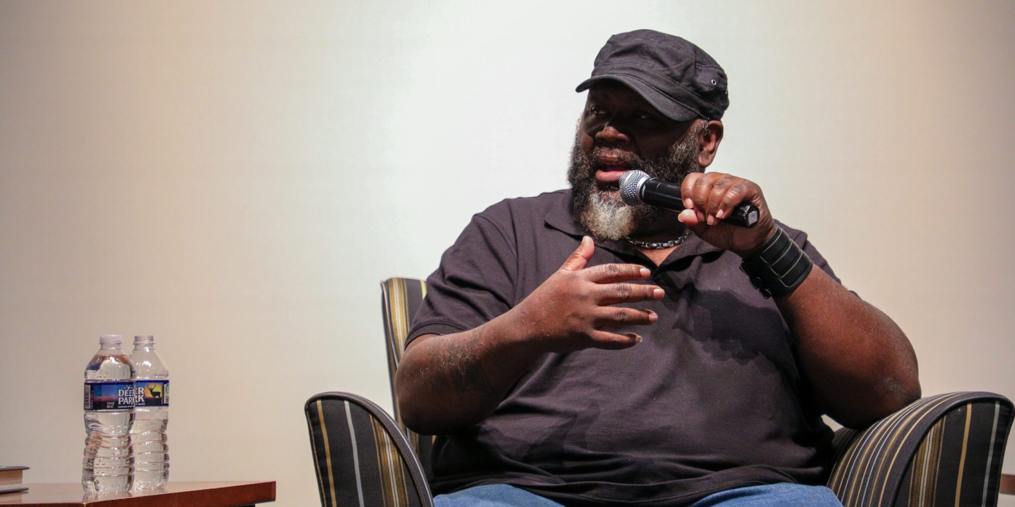 Author S.A. Cosby is seated in a chair, speaking into a microphone with two water bottles on a side table next to him