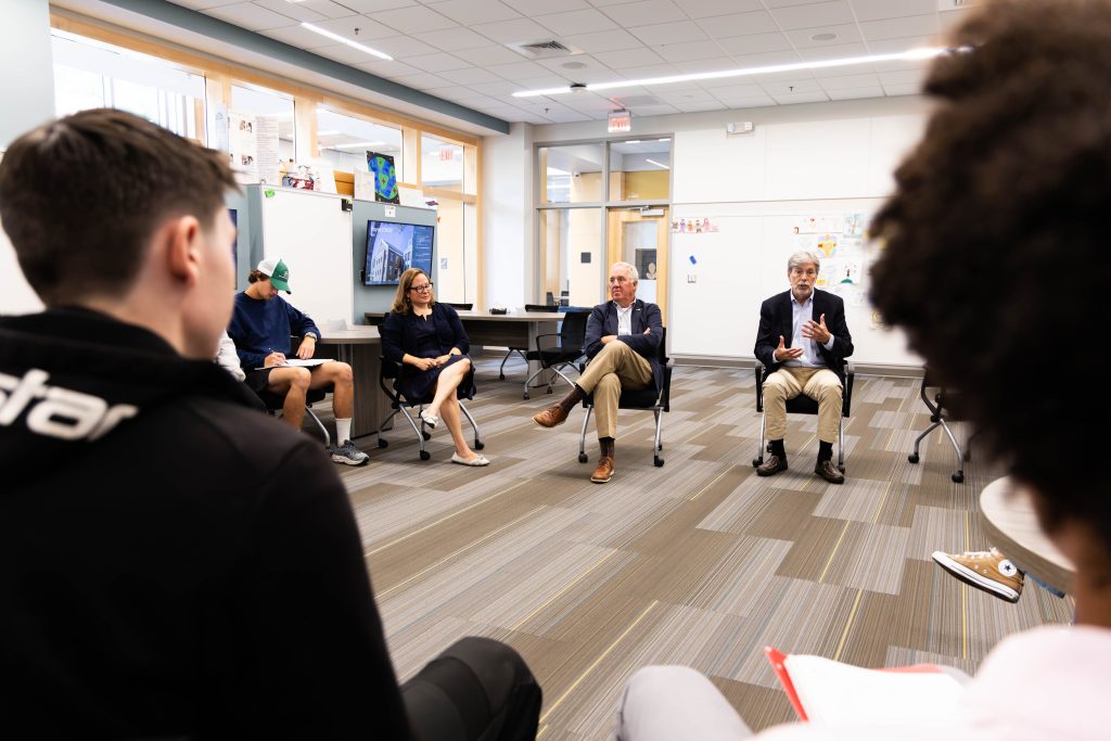 Two men in khakis and blazers talk to a circle of students taking notes inside a classroom