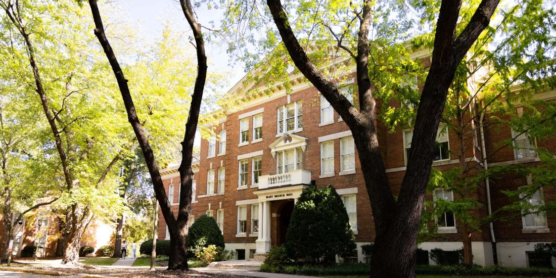 Large trees with green leaves provide shade in front of a large brick residence hall