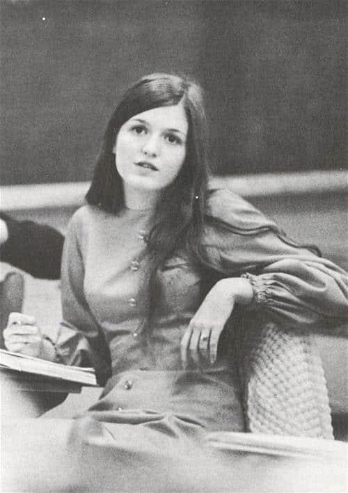 A woman in the 1970s sits at a desk in a college classroom.