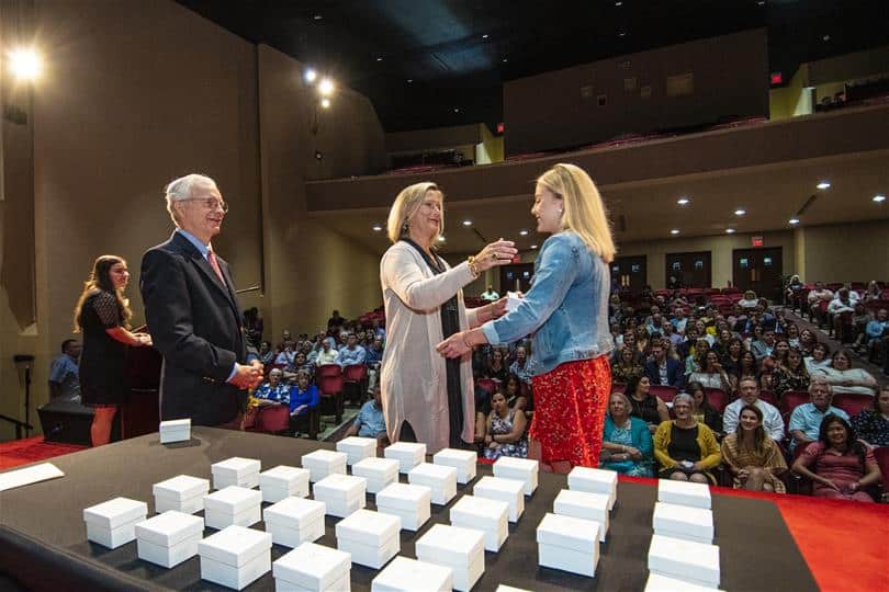 A shot from the stage of the Ring Ceremony. President Lindgren watches as a mother and daughter hug in front of a table filled with boxes of rings.